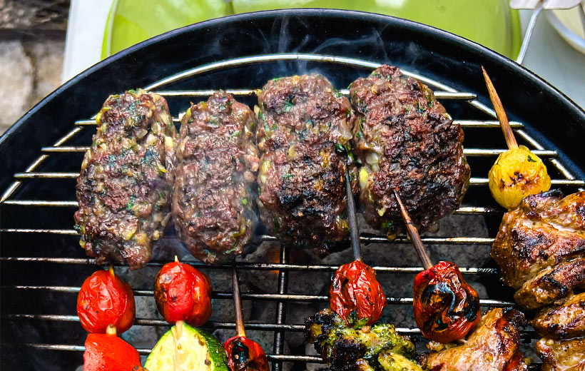 Hot Charcoal grill with four kebabs made of ground meat, chopped herbs, and Mediterranean spice blend "Kababalicious". Next to them on the grill are skewers with other grilled meat and grape tomatoes.