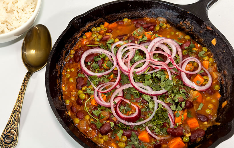 A cast iron pan with Rajma dish cooked with our Indian spice blend "Chili con Rajma". Cooked with kidney beans and other vegetables, and garnished with chopped cilantro and pickled red onion.