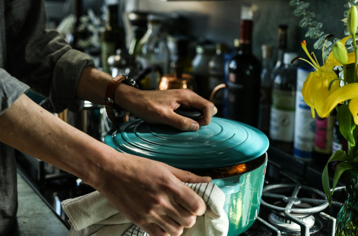 Hands of a cook holding a beautiful light blue cast iron pot on the stove. Behind are various bottles of wine and sauces.