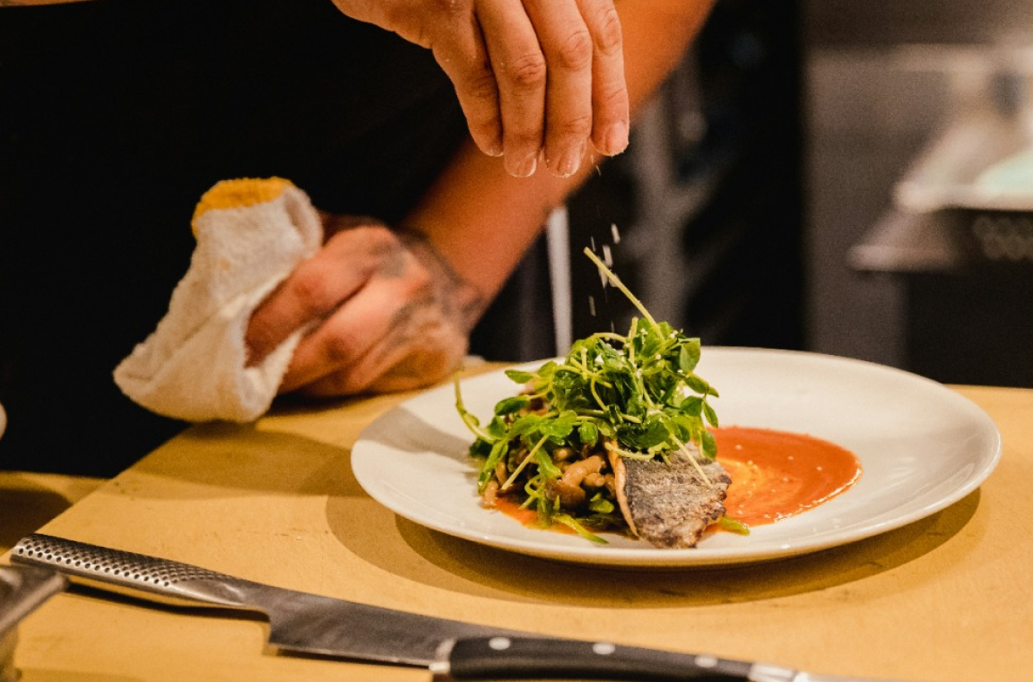 Chef's hand adding garnish to a plate with seared meat and salad topped over a splash of red sauce.