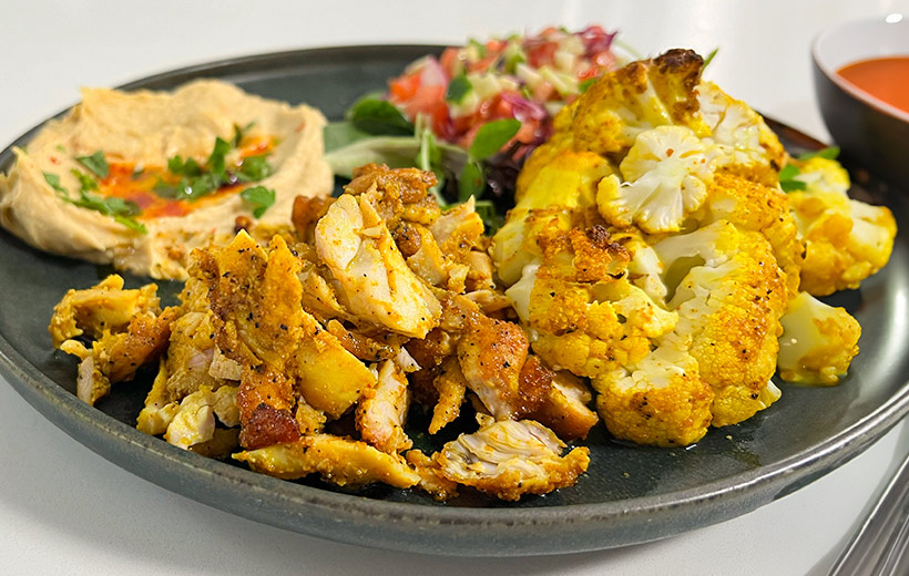 A plate of Mediterranean food with chicken shawarma and roasted cauliflower made with "Mama Shawarma" spice blend. Next to it hummus with paprika and garnish, and finely chopped salad.