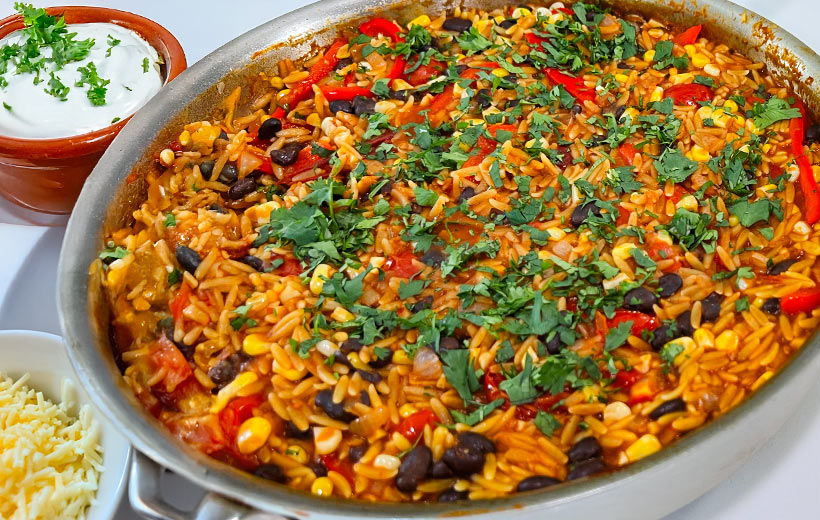 Stainless still pan with Mexican Orzo, with black beans, corn, red peppers and chopped cilantro. On the side, small serving bowls with sour cream and grated cheese.