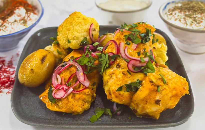 Dinner plate with beautiful chicken tikka garnished with chopped herbs and pickled red onion. On the side, roasted potatoes and cauliflower with cumin seeds.