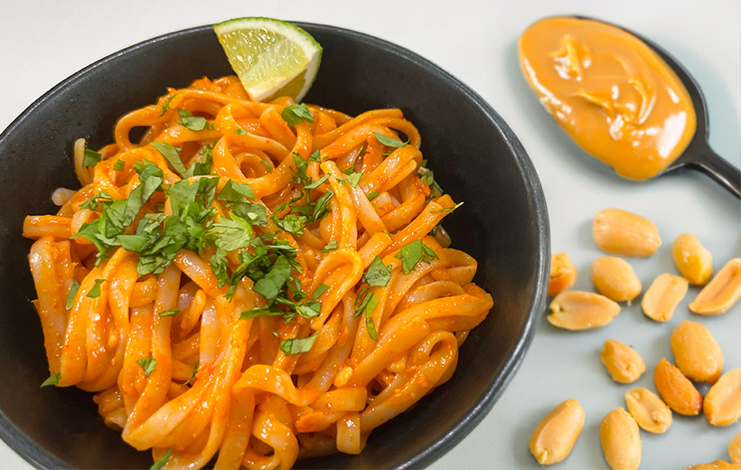 A black bowl with rice noodles in peanut butter sauce. garnished with chopped herb and lime wedge. Next a spoon with peanut butter, and peanuts scattered around it.