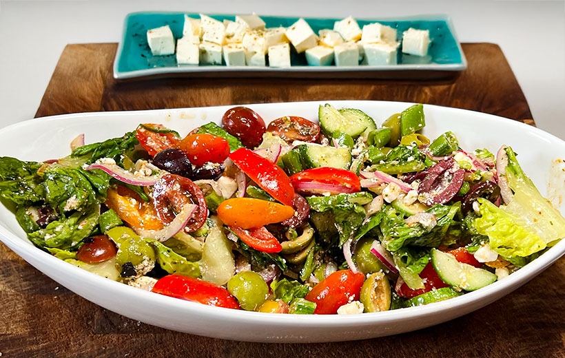 An oval white bowl with amazing looking greek salad including lettuce, cut tomatoes, red onion, kalamata olives mixed with vinaigrette with crumbled feta cheese. Behind, a turquoise place with cubed feta.