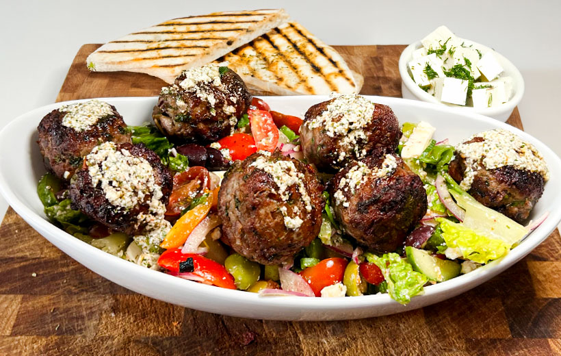 A bowl with beautiful lamb meatballs, over fresh greek salad, and topped with feta cheese vinaigrette. Behind it a small bowl with feta cheese, and some grill bread.