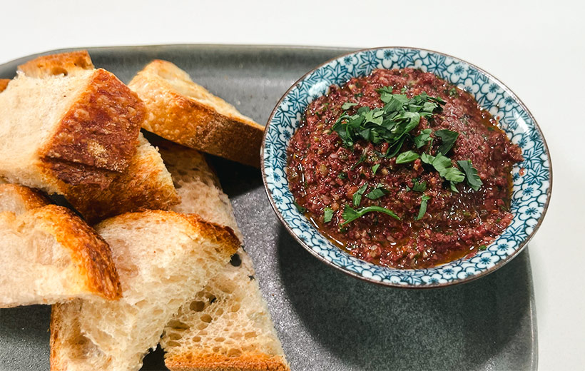 A gray dish with an ornamented bowl with fresh homemade olive tapenade and garnished with chopped parsley. Next, slice fresh sourdough bread